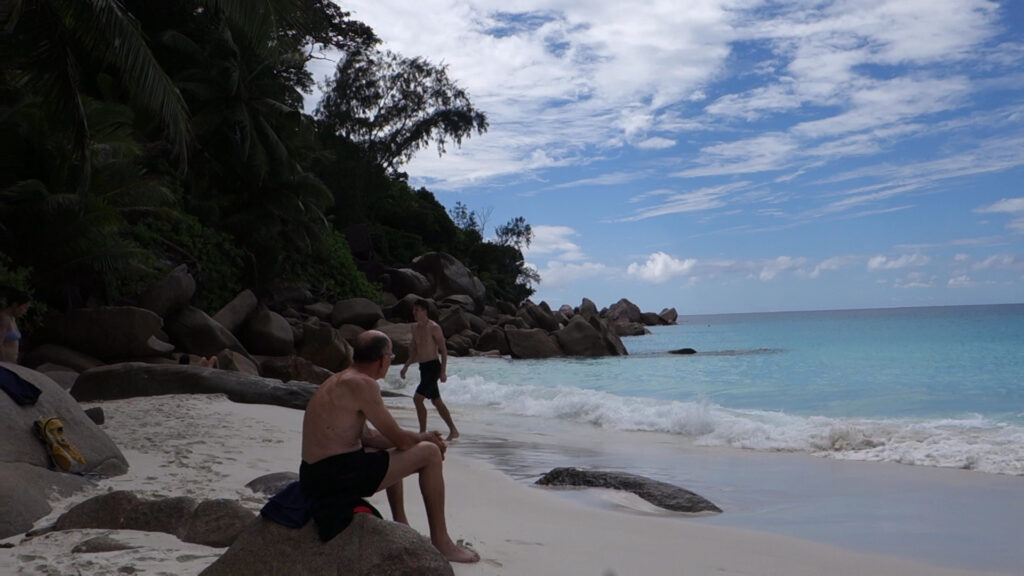 Zwei Männer am tropischen Strand; einer sitzt auf Felsen, der andere geht im seichten, türkisfarbenen Meer.