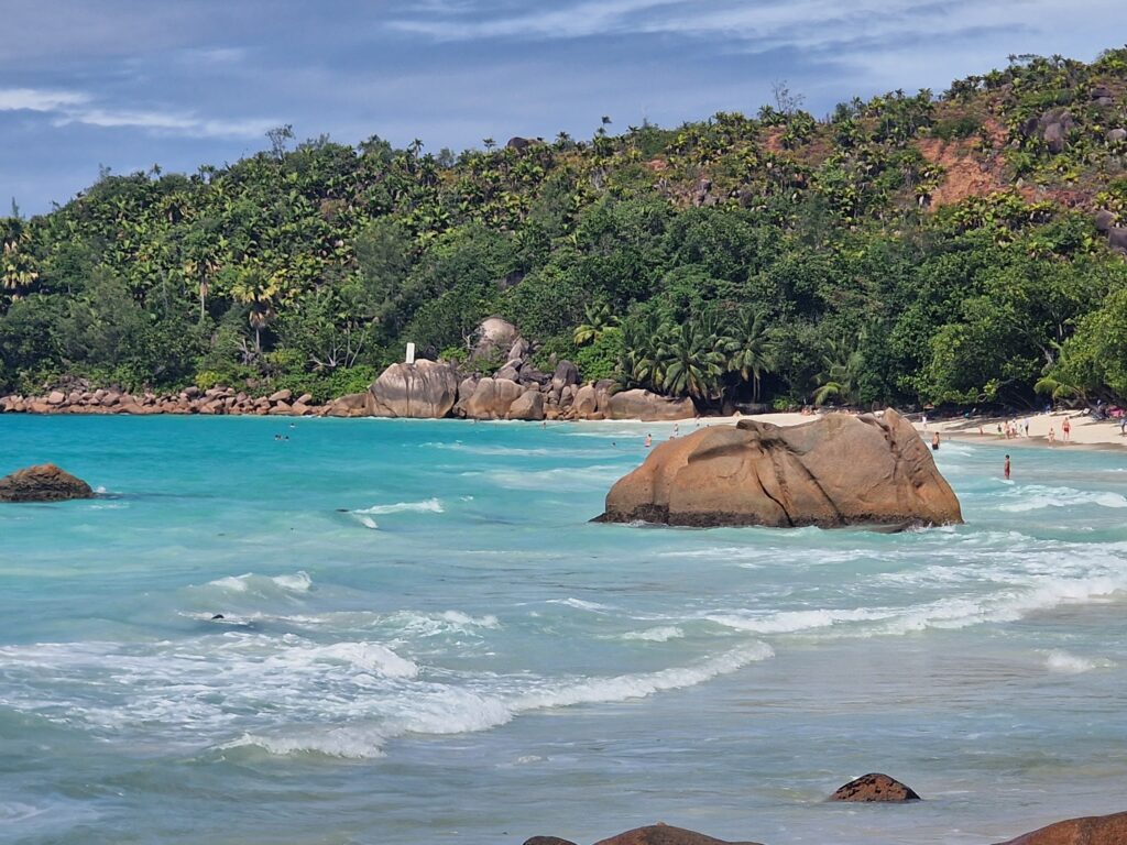 Türkisblaue Bucht mit Granitfelsen, palmengesäumtem Sandstrand und vereinzelten Badenden unter blauem Himmel.