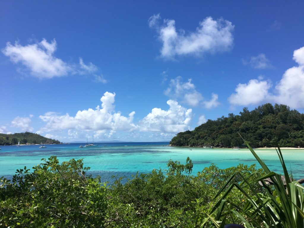 Türkisblaues Meer vor dicht bewaldeter Insel, klarer Himmel mit weißen Wolken, vereinzelte Segelboote in der Bucht.