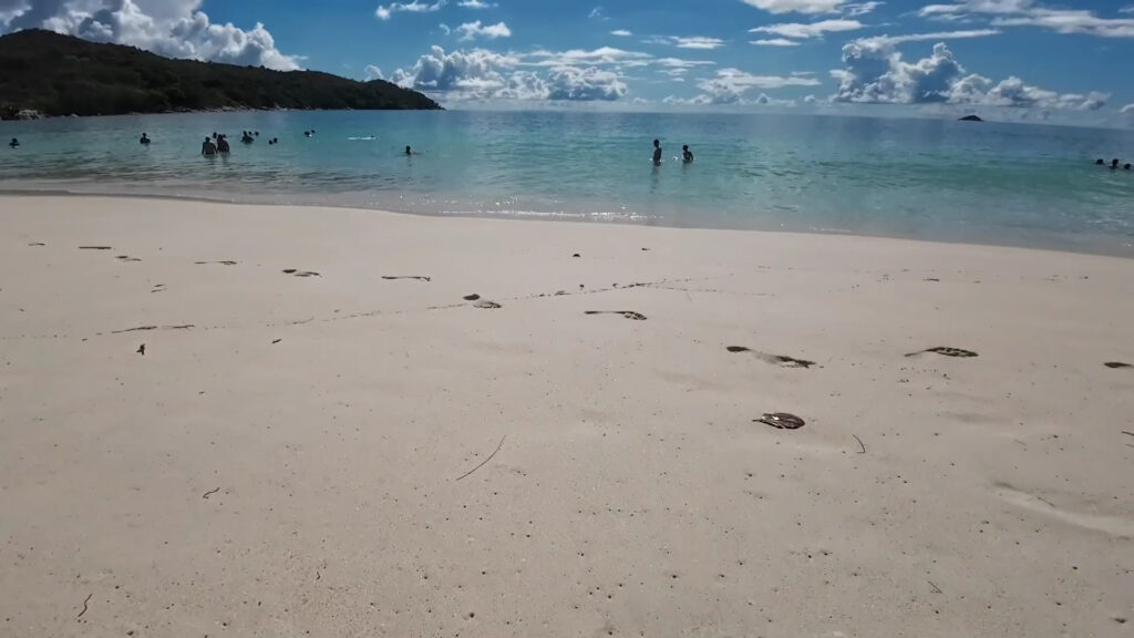 Heller Sandstrand mit Fußspuren, Menschen baden im klaren türkisfarbenen Meer vor bewaldeter Küste und Wolkenhimmel.