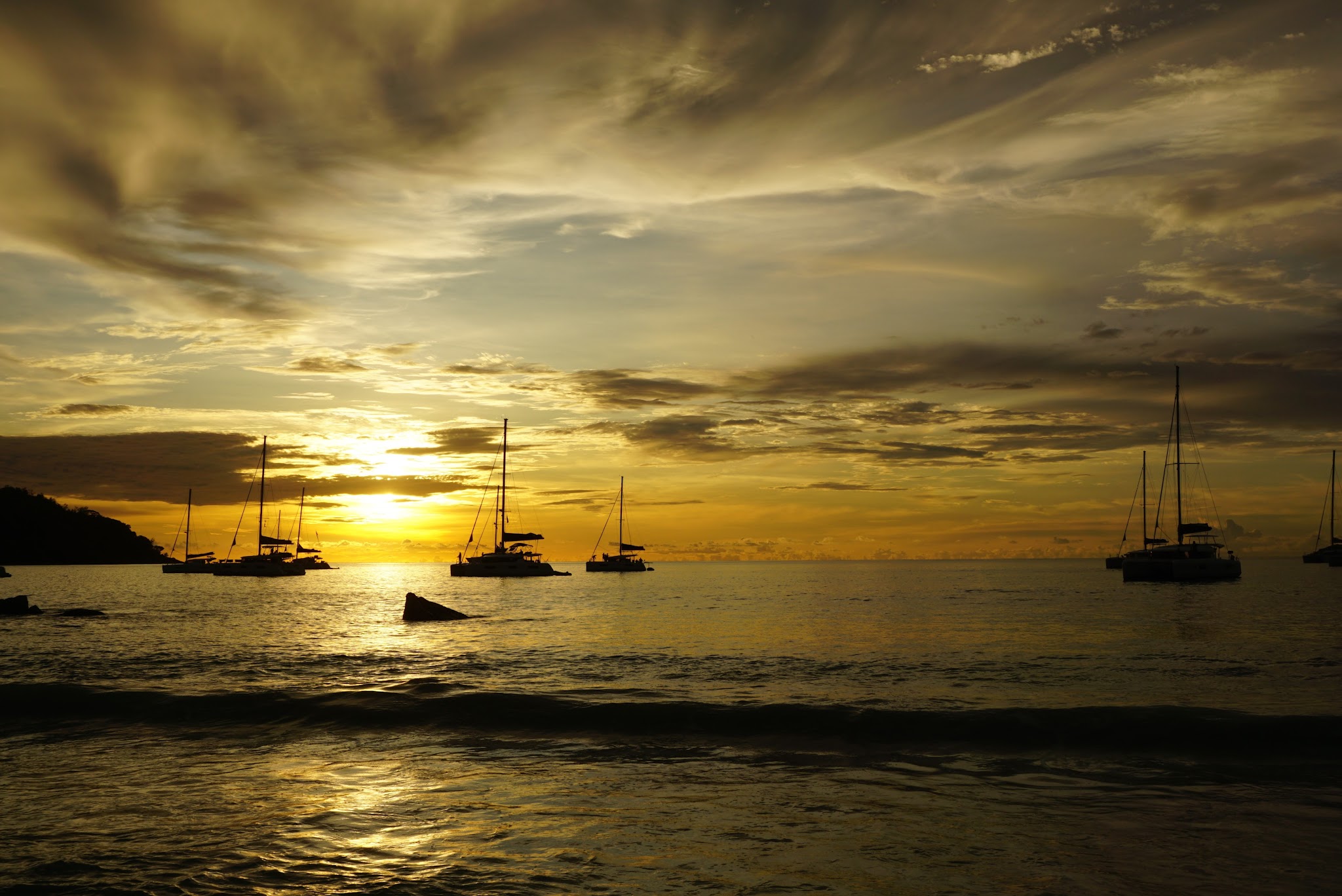 Mehrere Segelboote ankern im ruhigen Meer vor einem goldglühenden Sonnenuntergang unter dramatisch bewölktem Himmel