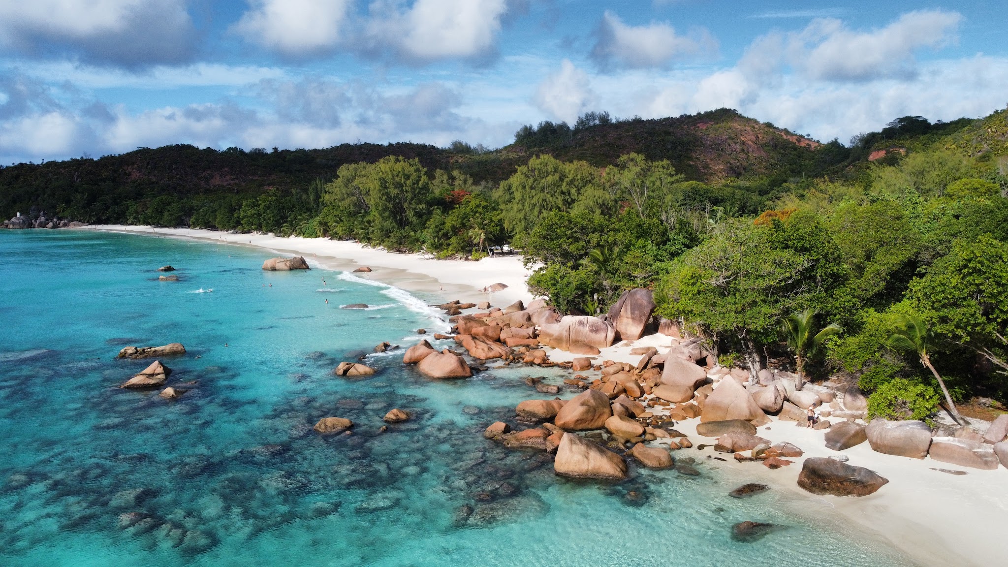 Türkisblaue Bucht mit weißem Sandstrand, runden Granitfelsen im Wasser und dichtem Wald bis zu den Hügeln.