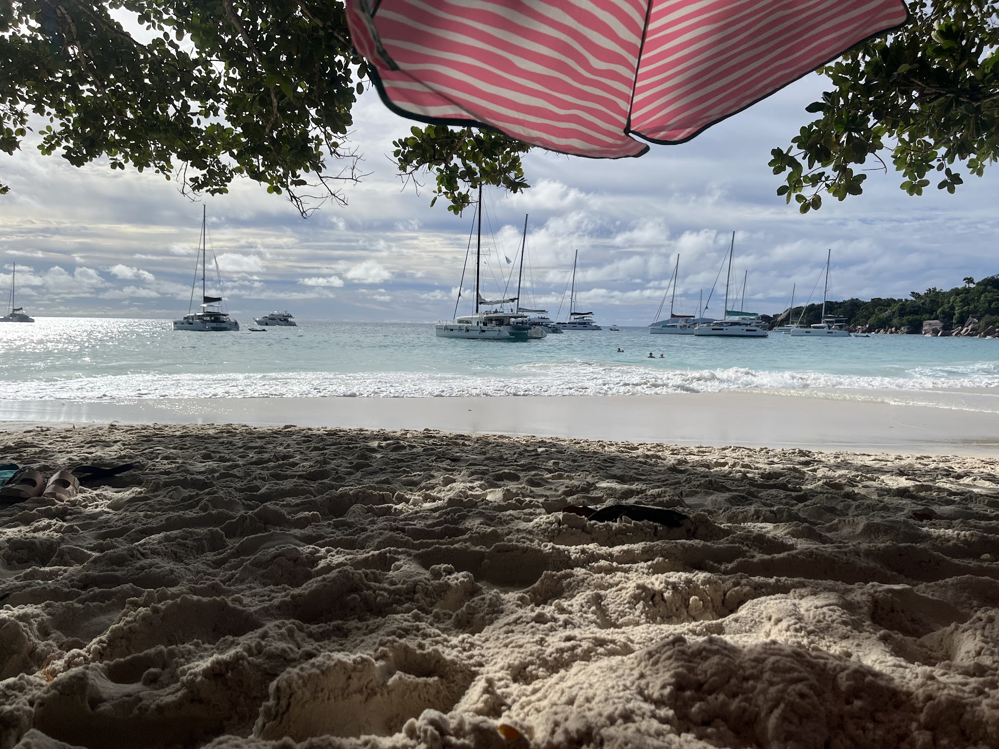 Sonnendurchfluteter Sandstrand unter rosa-weißem Schirm, davor türkisfarbenes Meer mit mehreren Segelbooten.
