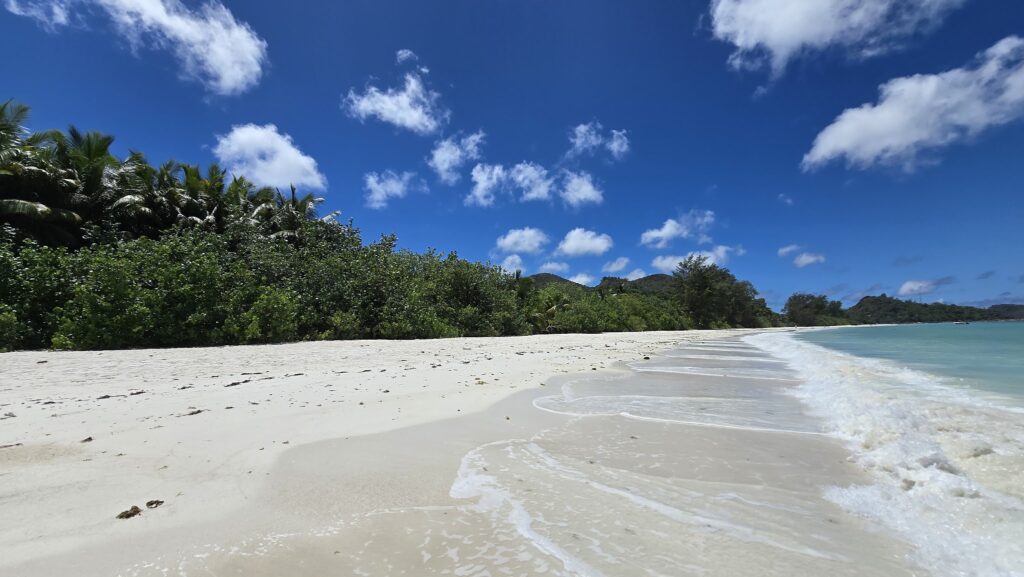 Weißer Sandstrand mit sanften Wellen, türkisfarbenem Meer, Palmen und blauem Himmel mit lockeren Wolken.