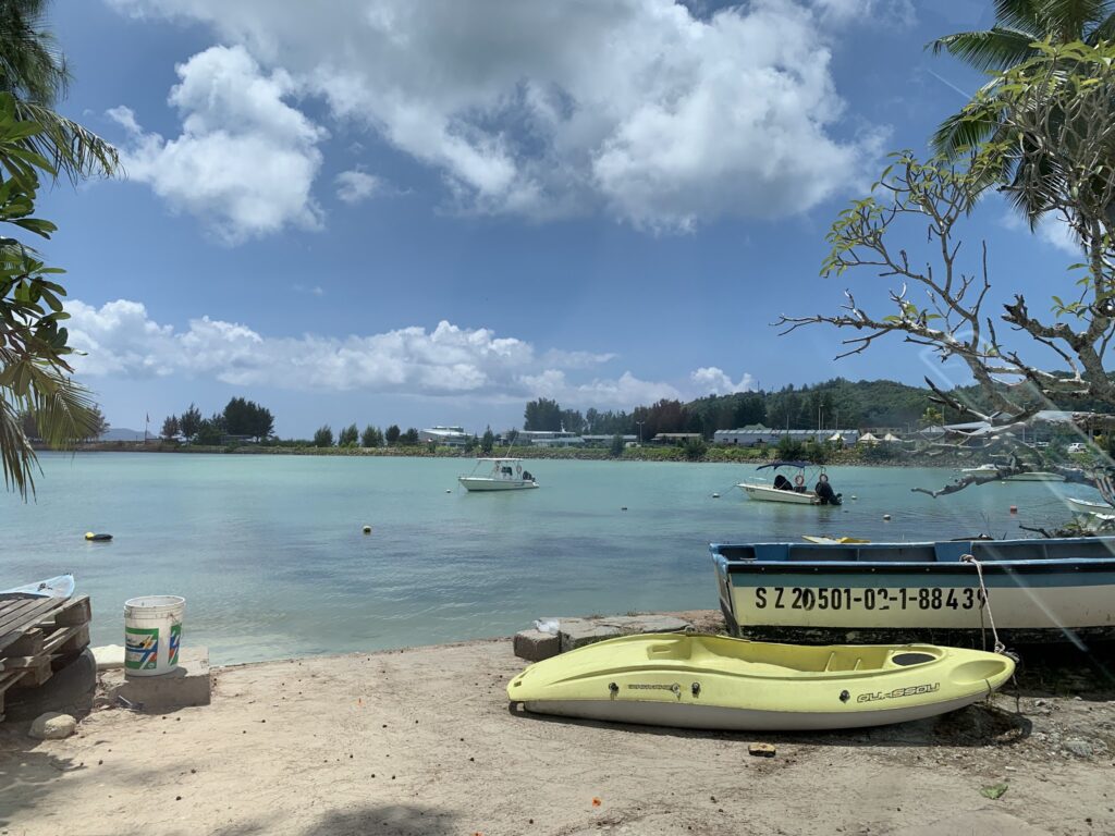 Türkisblaue Lagune mit kleinen Motorbooten; gelbes Kajak und Fischerboot liegen am sandigen Ufer, Palmen im Hintergrund.