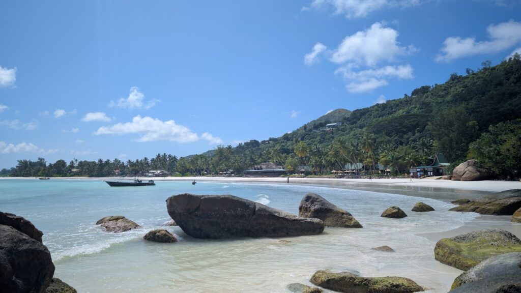 Türkisblaue Bucht mit Palmenstrand, Felsen im flachen Wasser, kleines Boot vor grün bewaldetem Hügel unter blauem Himmel