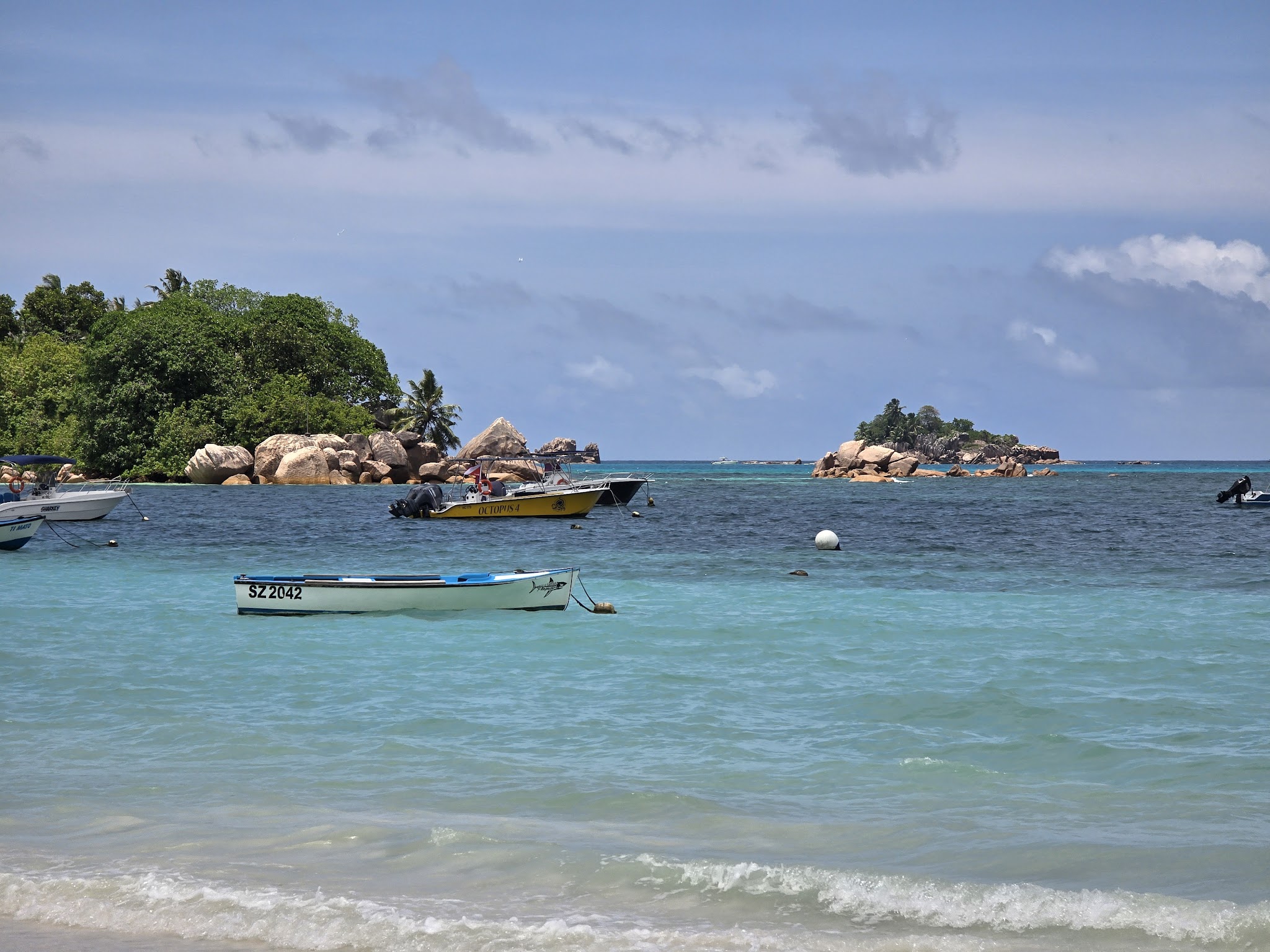 Türkises Meer mit drei kleinen Booten vor zwei palmengesäumten Granitinseln unter blauem Himmel.