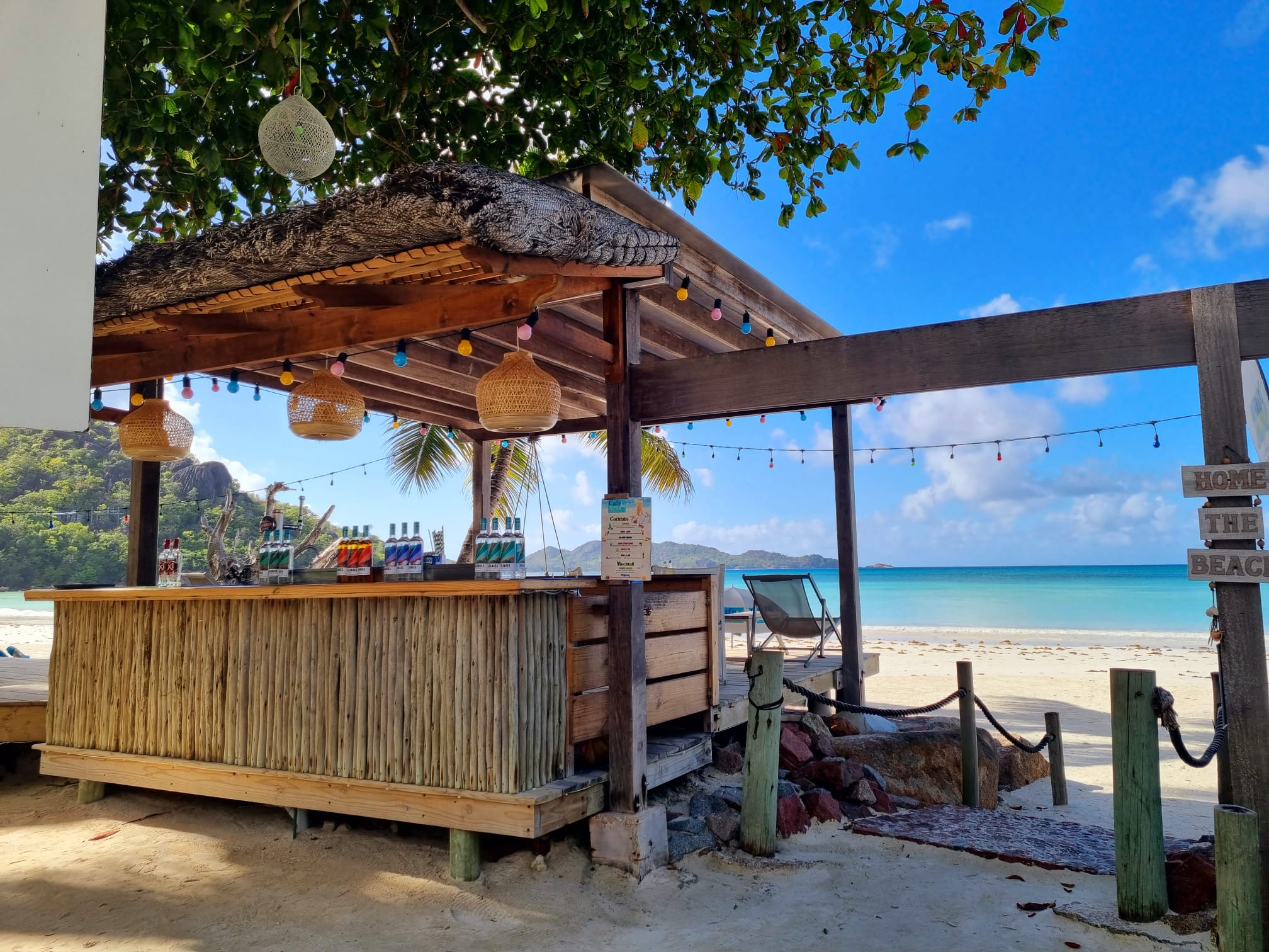 Offene Bambus-Strandbar mit bunten Spirituosenflaschen, Lampions und Lichterkette vor türkisblauem Meer und weißem Sand.