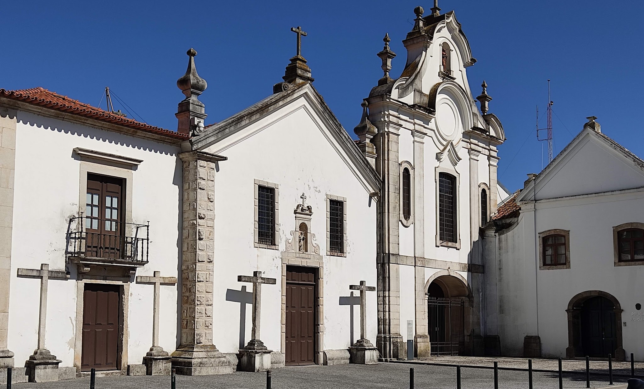 Barocke, weiß verputzte Kirchenfassade mit zwei Türmen und drei steinernen Kreuzen, davor sonniger Platz, blauer Himmel