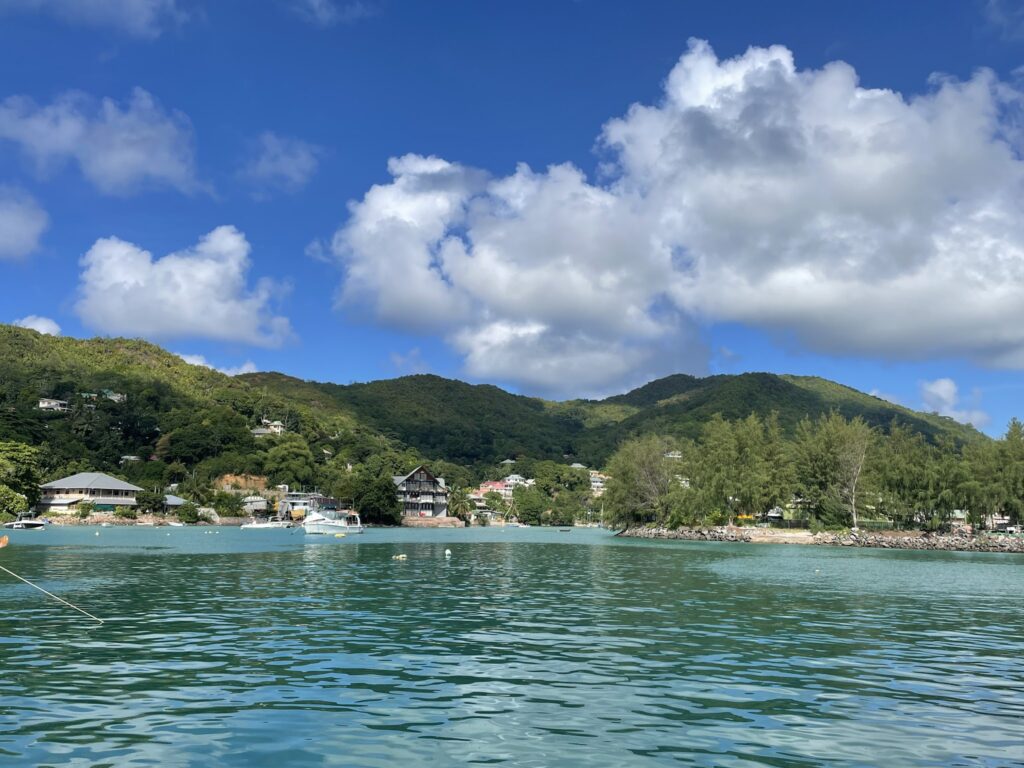 Türkisblaue Bucht mit kleinen Booten vor bewaldeten Hügeln, verstreuten Häusern und wolkigem, strahlend blauem Himmel.