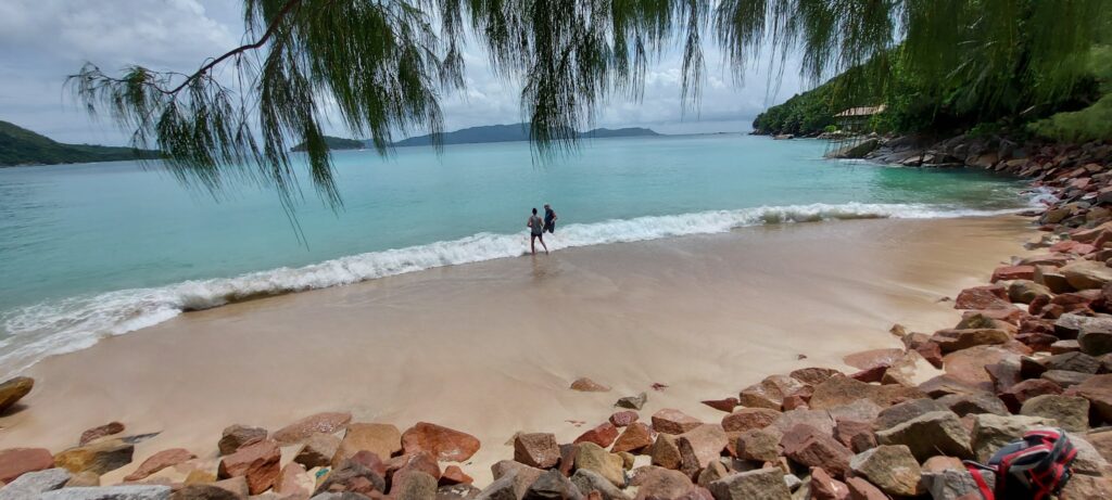 Zwei Menschen stehen am Rand türkisblauer Wellen an einer ruhigen, von Felsbrocken gesäumten Tropenbucht.