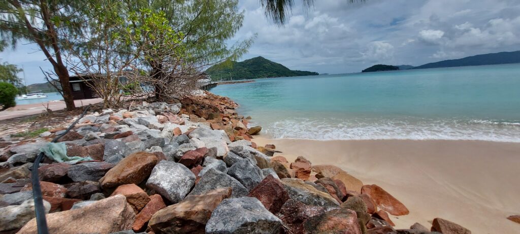 Felsige Uferböschung neben sandigem Strand, türkisblaues Meer mit grünen Hügeln unter teils bewölktem Himmel