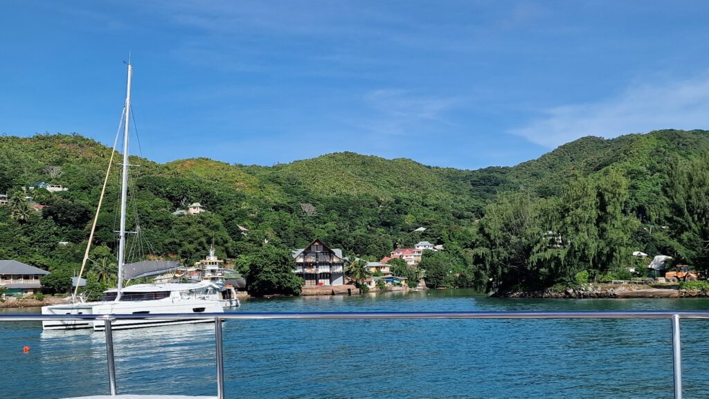 Weißes Segelkatamaran vor grünen Hügeln und bunten Häusern, gesehen hinter Reling über ruhiger Bucht