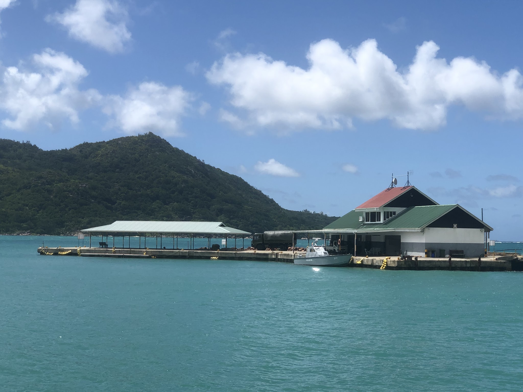 Kleiner Pier mit Bootshaus und Boot im türkisfarbenen Meer, grüner Inselberg und blauer Himmel im Hintergrund.
