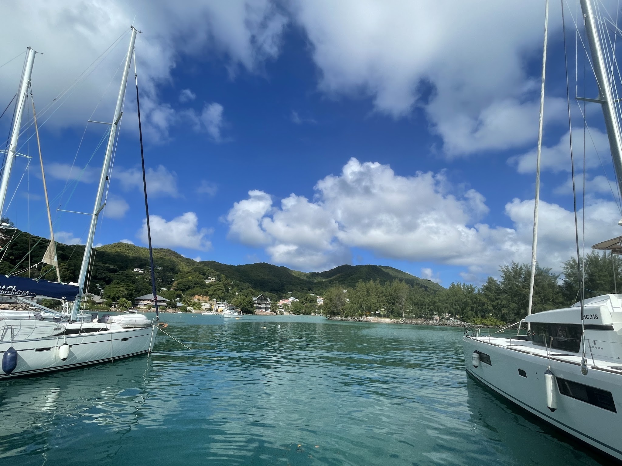 Zwei Segelboote rahmen eine türkisfarbene Bucht vor grünen Hügeln und blauem Himmel mit weißen Wolken.