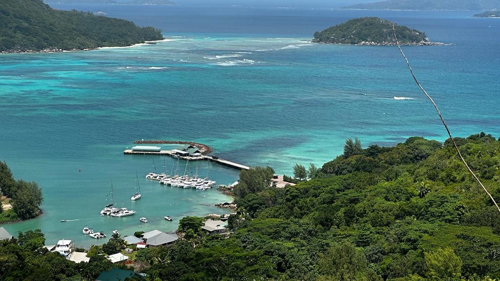Türkisfarbenes Meer mit kleiner Marina voller Segelboote, umgeben von dichtem Grün, vorgelagerten Inseln im Hintergrund.