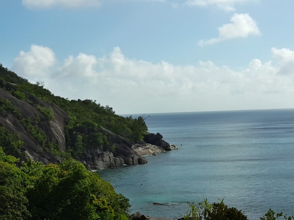 Granitklippen mit üppigem Grün ragen in ruhiges türkisblaues Meer unter leicht bewölktem Himmel.