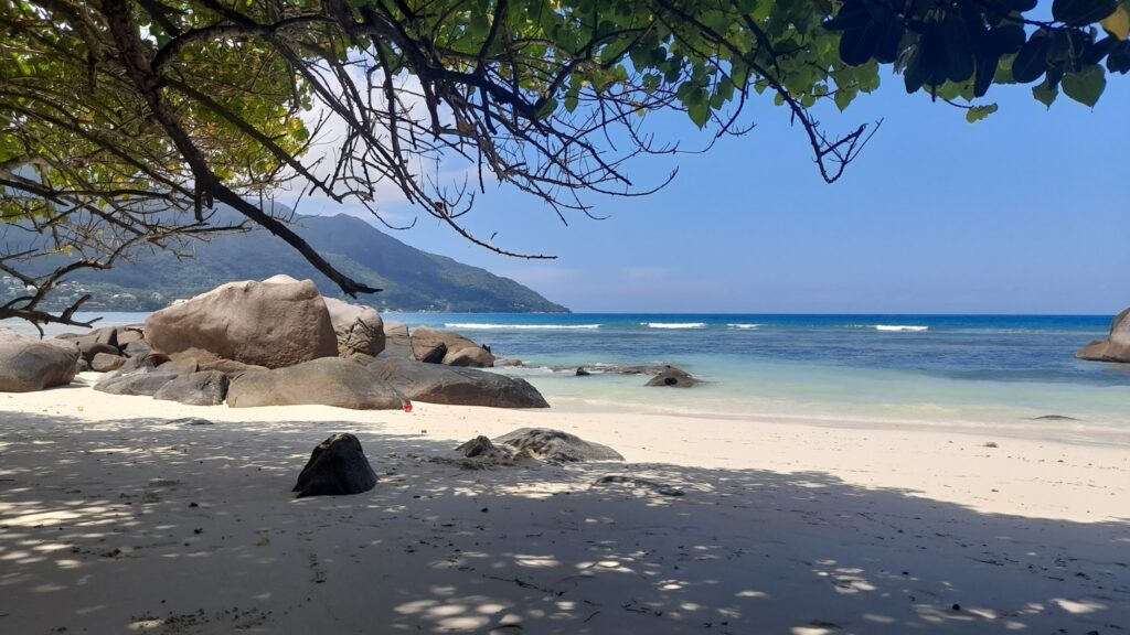 Verschatteter Sandstrand mit Granitfelsen, türkisblauem Meer und bewaldetem Berg im Hintergrund.