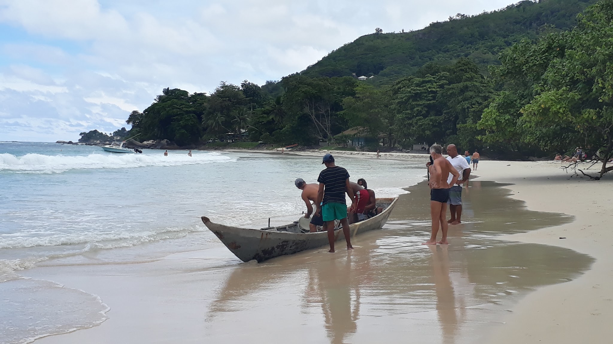 Mehrere Männer ziehen ein kleines Boot am tropischen Sandstrand an Land; Wellen und bewaldeter Hügel im Hintergrund.