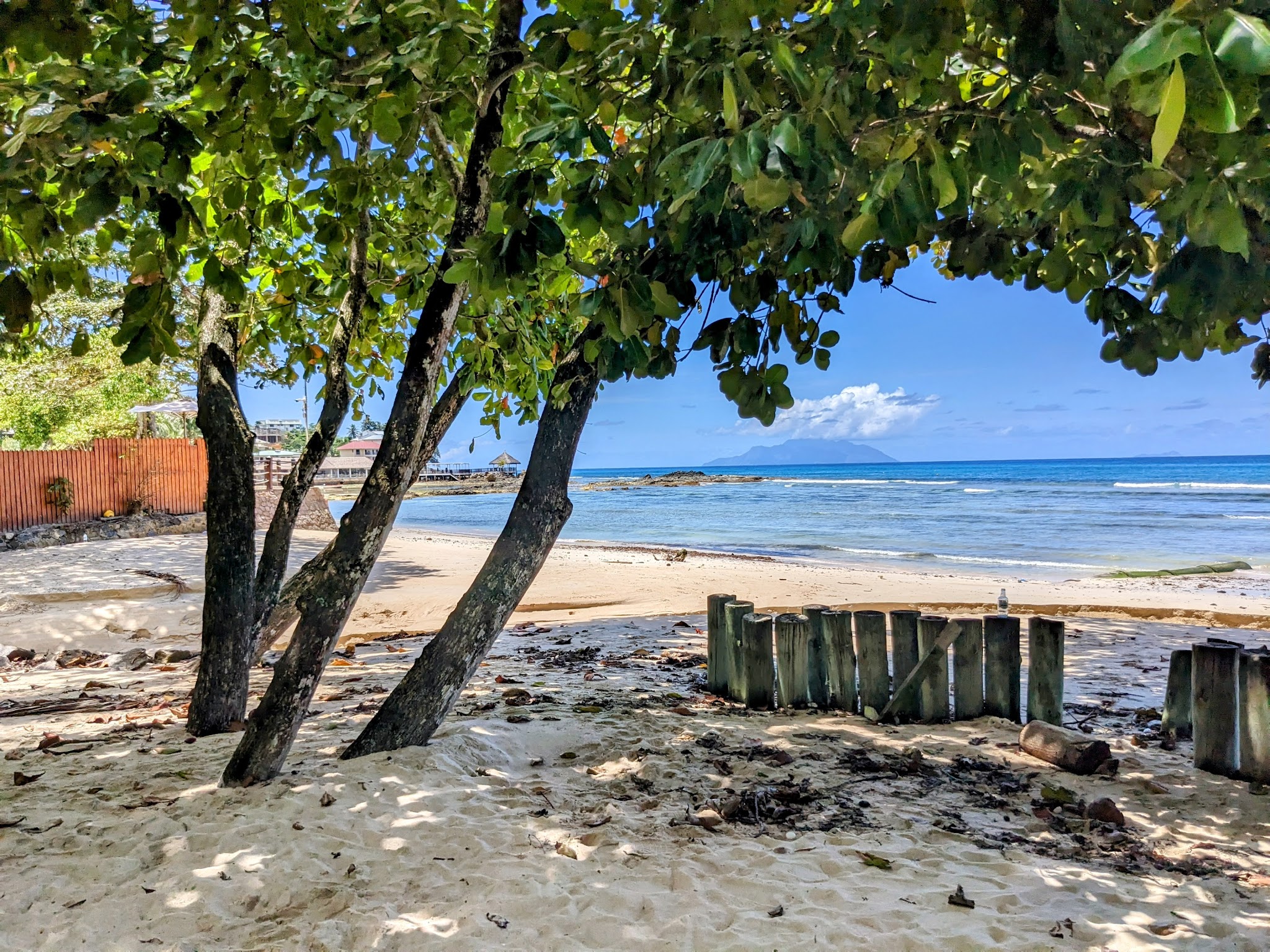 Schattige Bäume am Sandstrand, türkisblaues Meer mit sanften Wellen, ferne Vulkaninsel und kleine Steganlage.