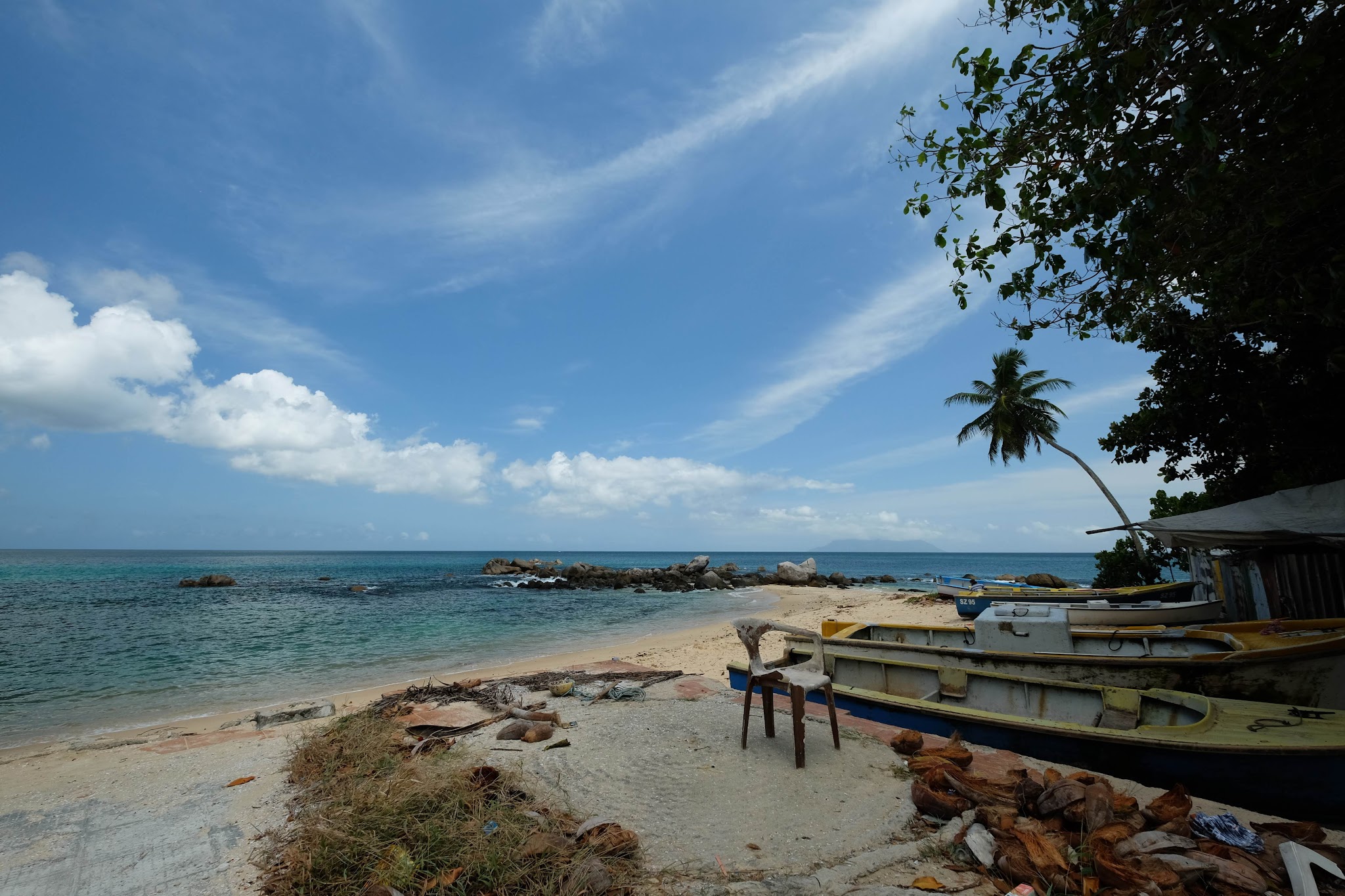 Strand mit türkisfarbenem Meer, einigen Fischerbooten, einsamer Plastiksessel im Sand, schräg wachsender Palme.
