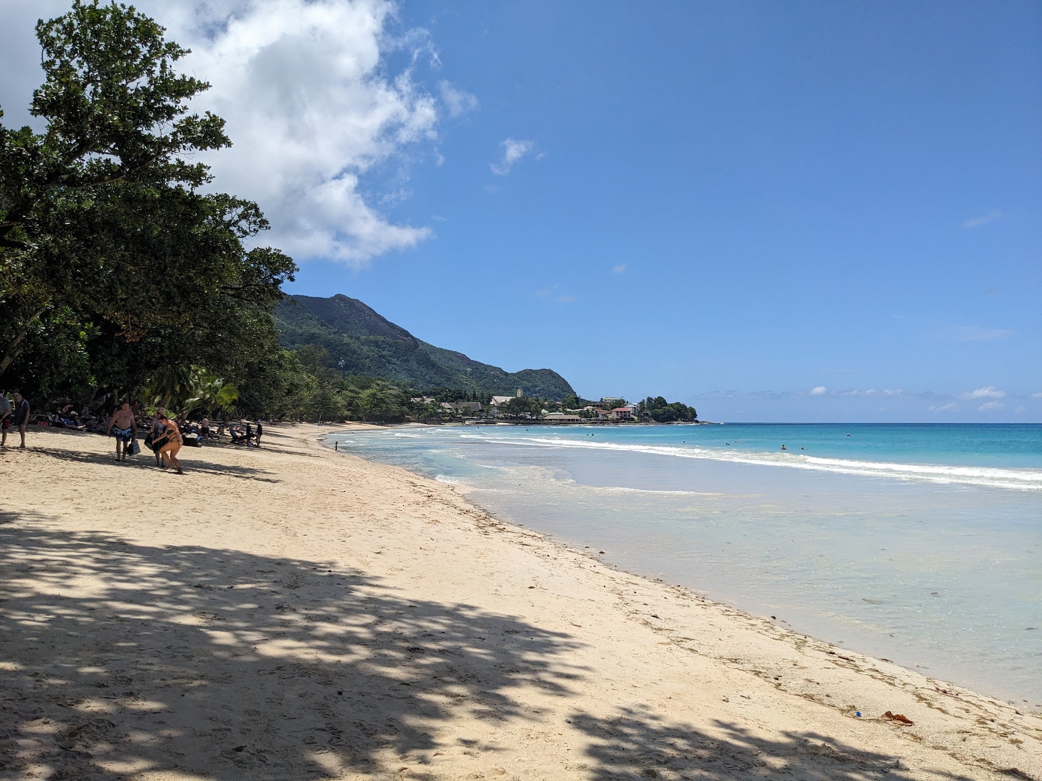 Breiter Sandstrand mit Schatten von Bäumen, türkisfarbenes Meer, im Hintergrund grüne Berge und wenige Badende