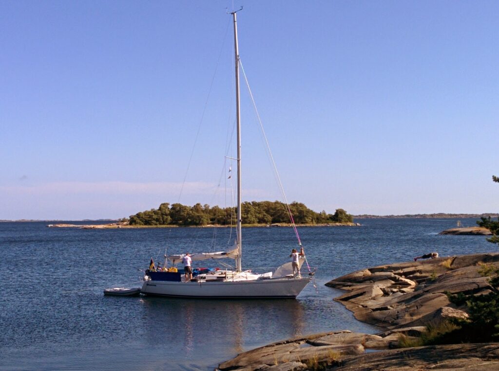 Segelboot ankert an felsiger Küste; zwei Personen an Bord, bewaldete Insel im ruhigen blauen Wasser dahinter.