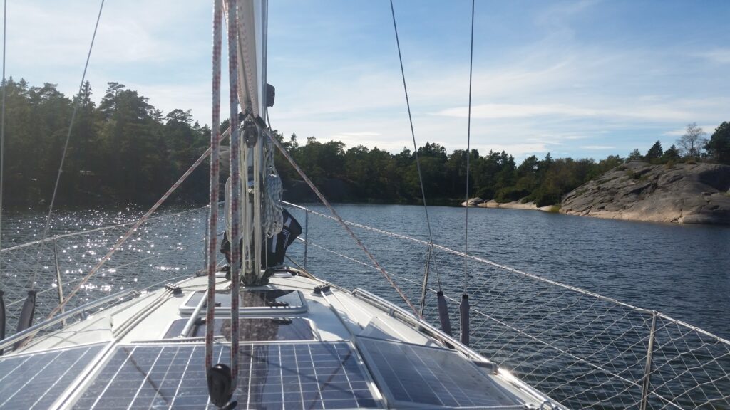 Blick vom Segelbootdeck mit Solarpaneelen über glitzerndes Wasser auf bewaldete Felsküste unter blauem Himmel.