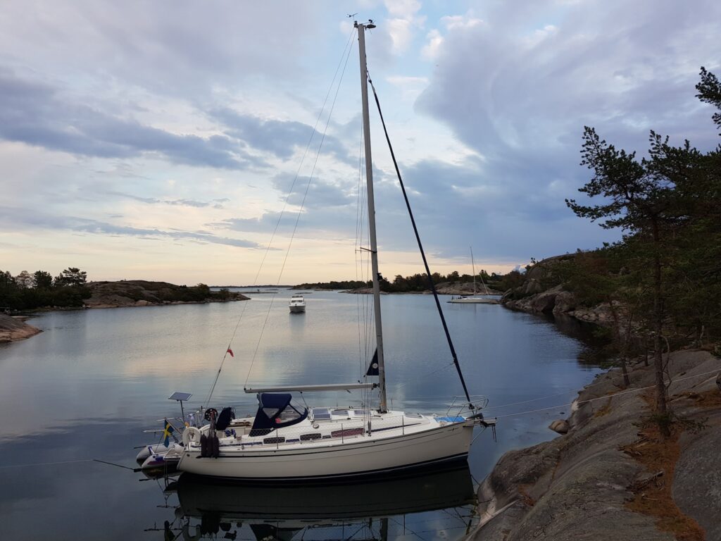 Segelboot vertäut in ruhiger, felsiger Bucht, sanfter Abendhimmel mit Wolken und fernen Booten.