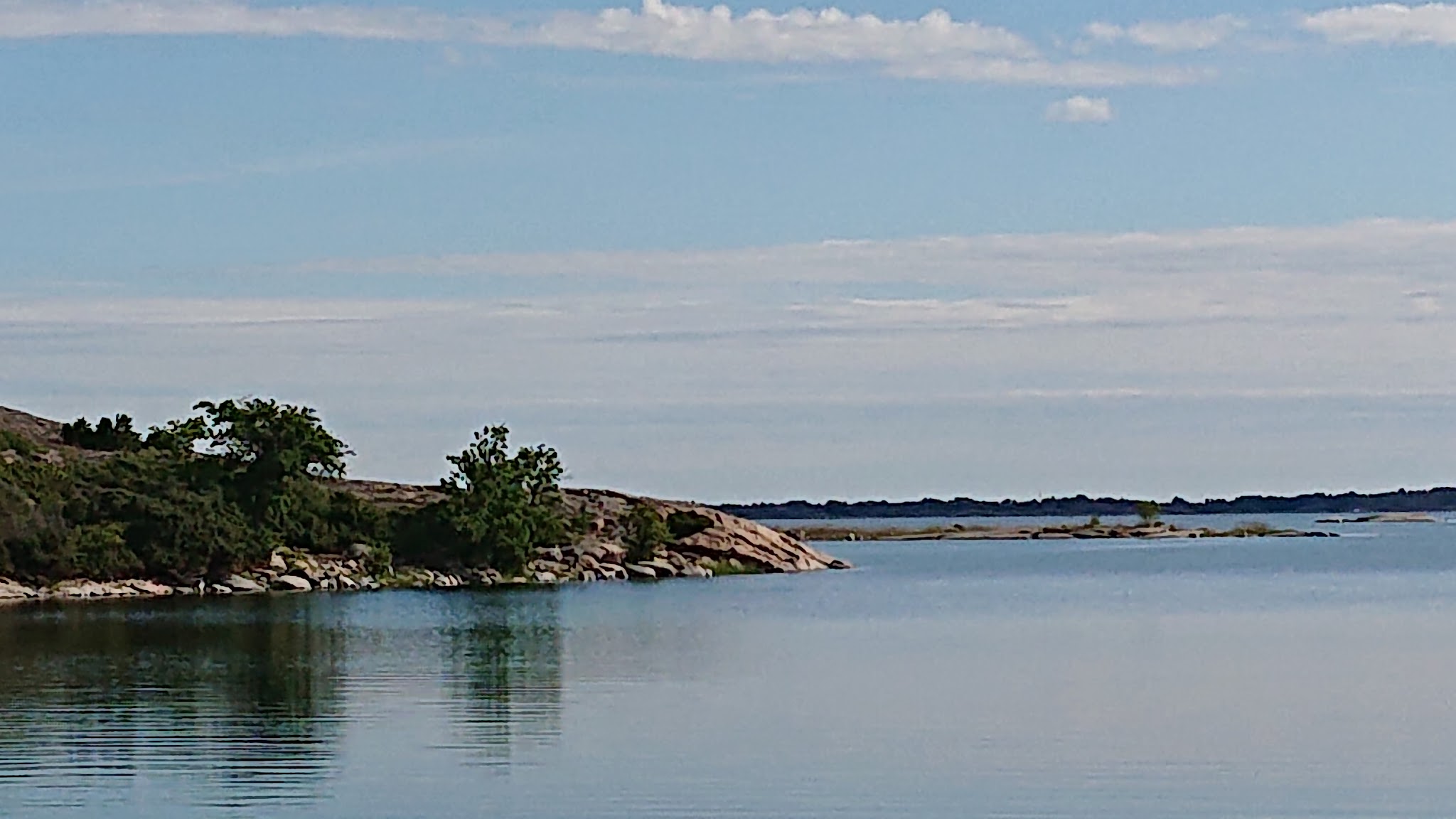 Ruhiges Küstenpanorama: glattes Wasser, kleine bewaldete Felsinsel links, ferner Horizont und leichter, wolkiger Himmel