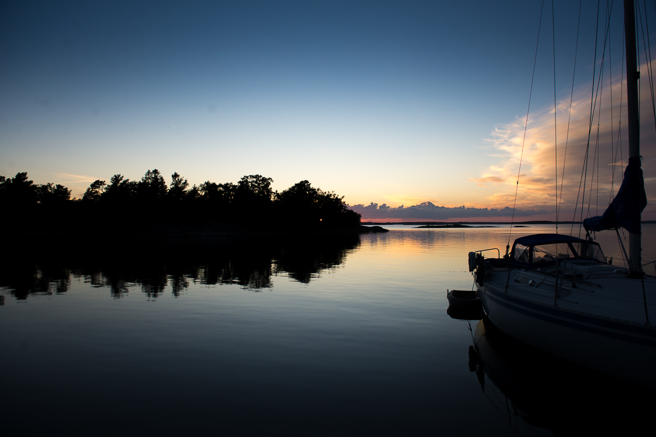 Ruhiger See bei Sonnenuntergang, dunkle Baumreihe spiegelt sich im Wasser, Segelboot liegt rechts im Vordergrund.