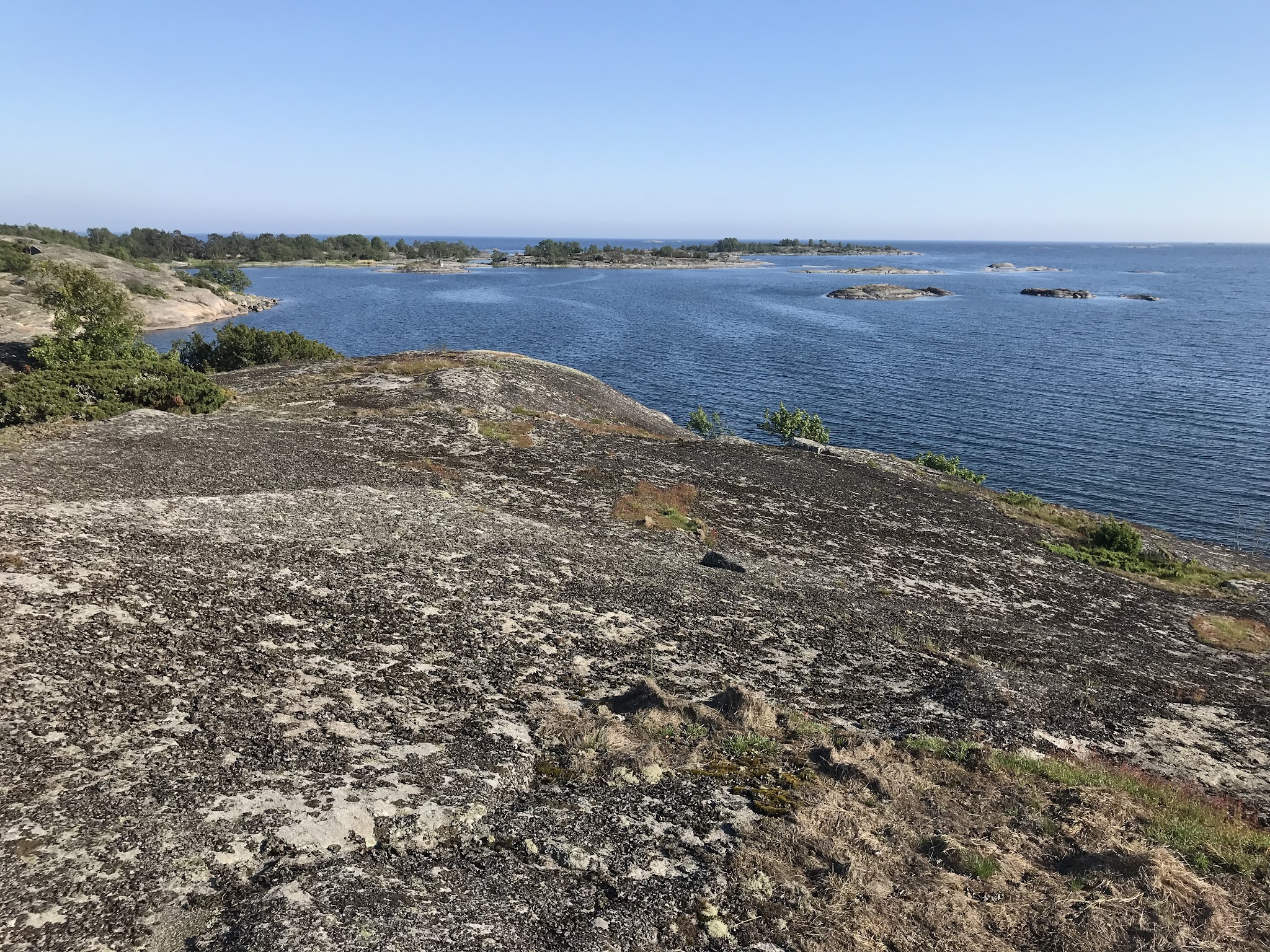 Karger Felsen mit Flechten am Meer, dahinter blaues Wasser mit kleinen Inseln und klarer Himmel