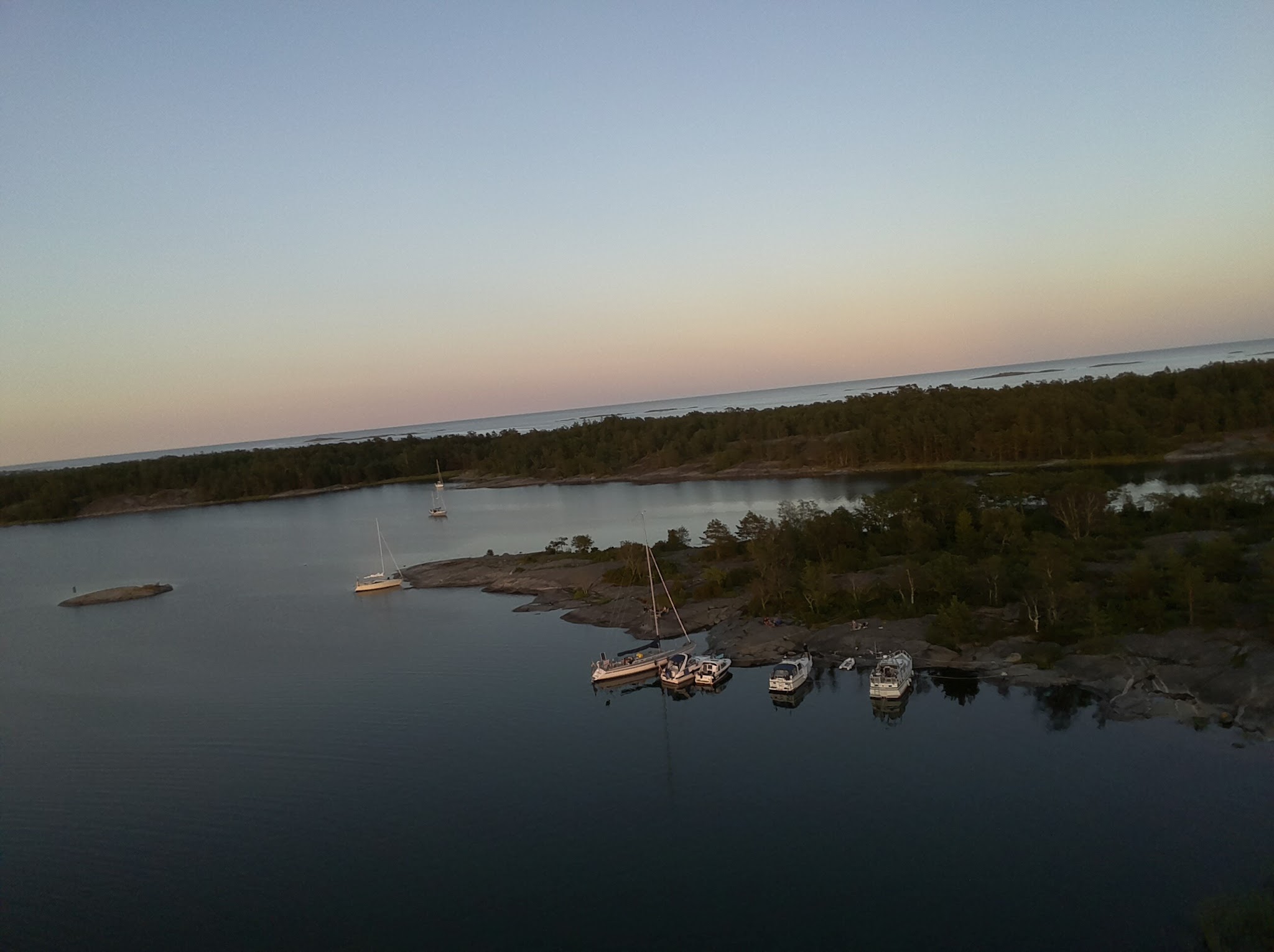 Segel- und Motorboote ankern in ruhiger Schärenbucht; Waldküste und pastellfarbener Sonnenuntergangshimmel.