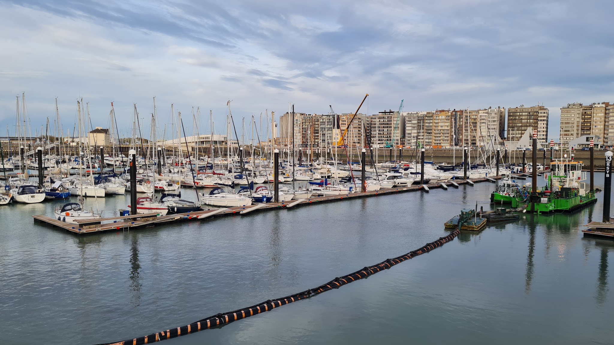 Yachthafen voller Segelboote vor einer Reihe hoher Wohngebäude unter leicht bewölktem Himmel