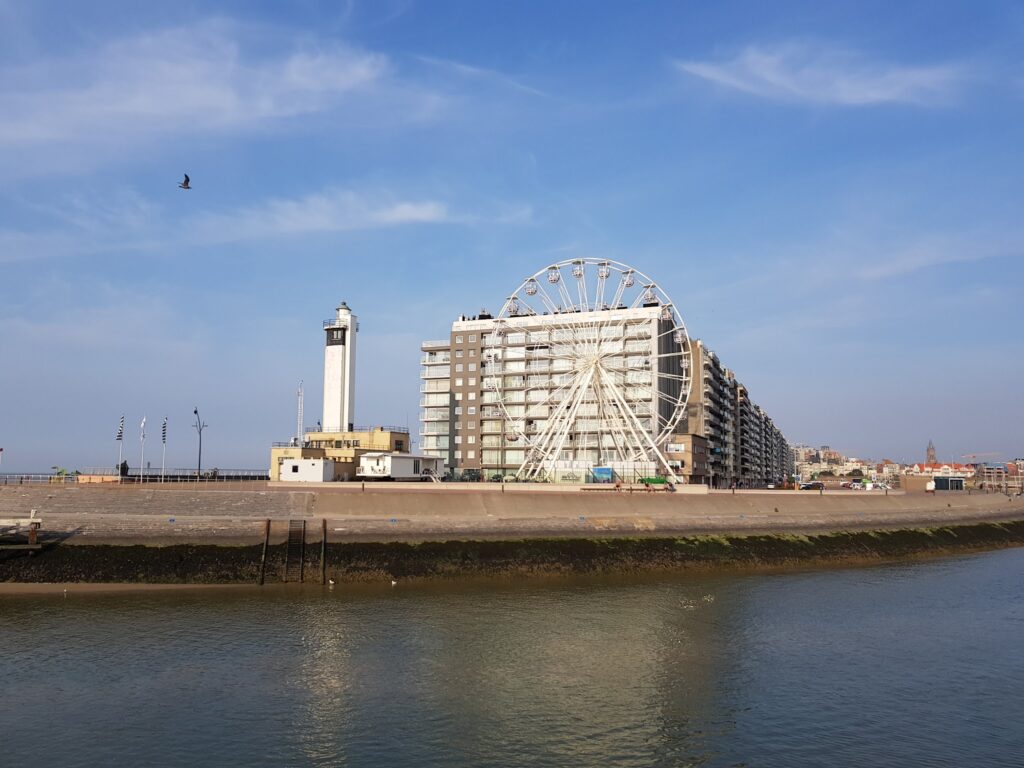 Seepromenade mit hohem Wohnblock, weißem Riesenrad und Leuchtturm am ruhigen Wasser unter blauem Himmel
