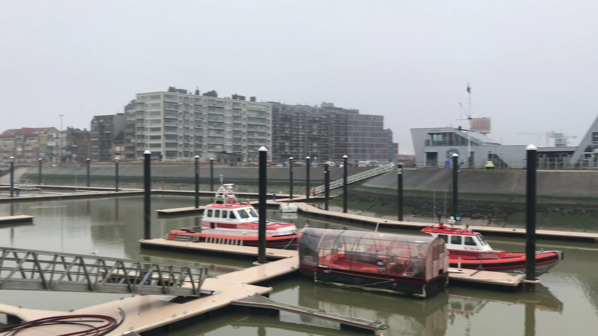 Kleiner Hafen; mehrere rot-weiße Rettungsboote liegen an schwimmenden Stegen vor grauen Wohnblöcken im Dunst.