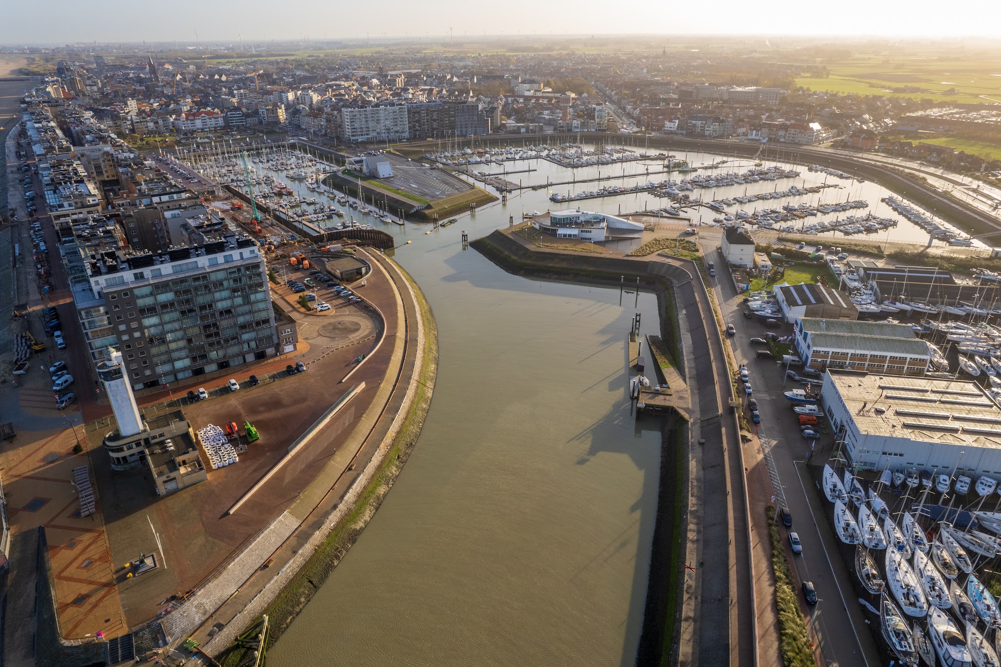 Luftaufnahme eines Küstenhafens mit Marina, dicht liegenden Segelbooten, Kanaleinfahrt und angrenzender Stadtbebauung.