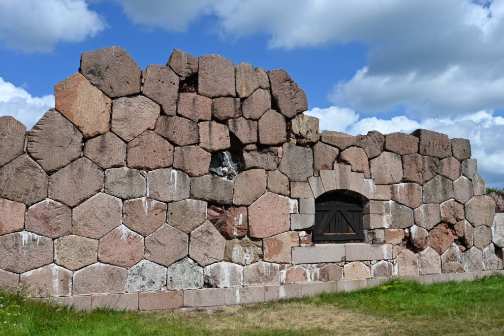 Mauer aus großen, unregelmäßigen sechseckigen Steinen mit kleinem, rundbogigem Holzfenster unter blauem Himmel