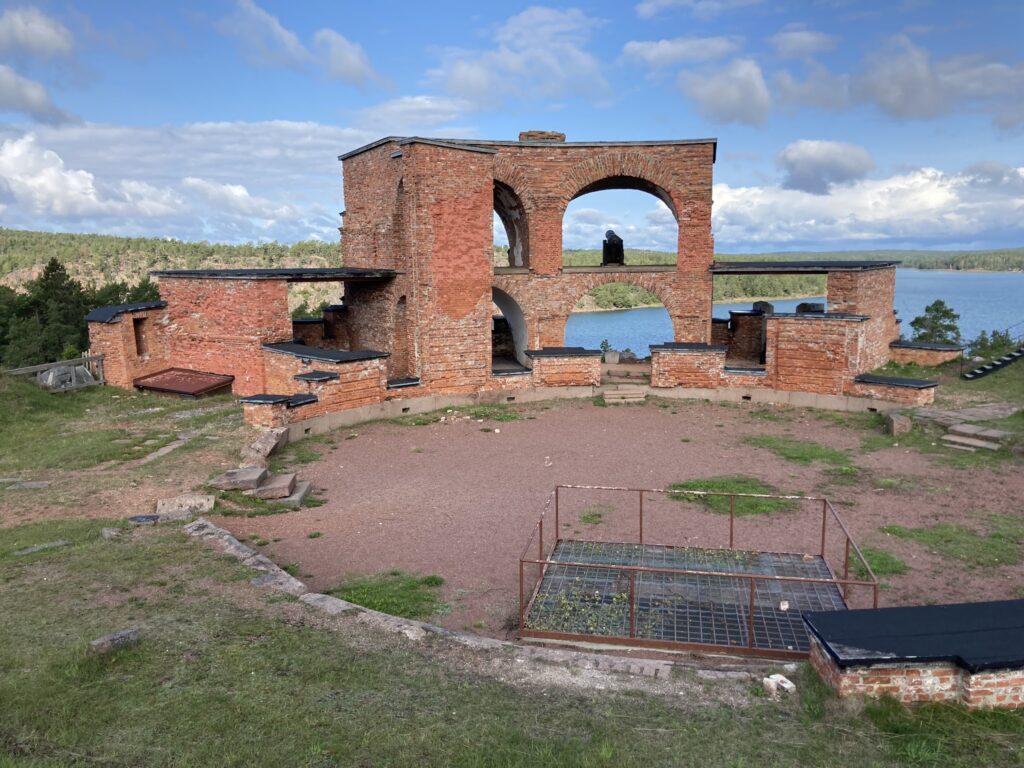 Backsteinruine mit großen Rundbögen auf Hügel, dahinter Wald und blauer See unter teils bewölktem Himmel.