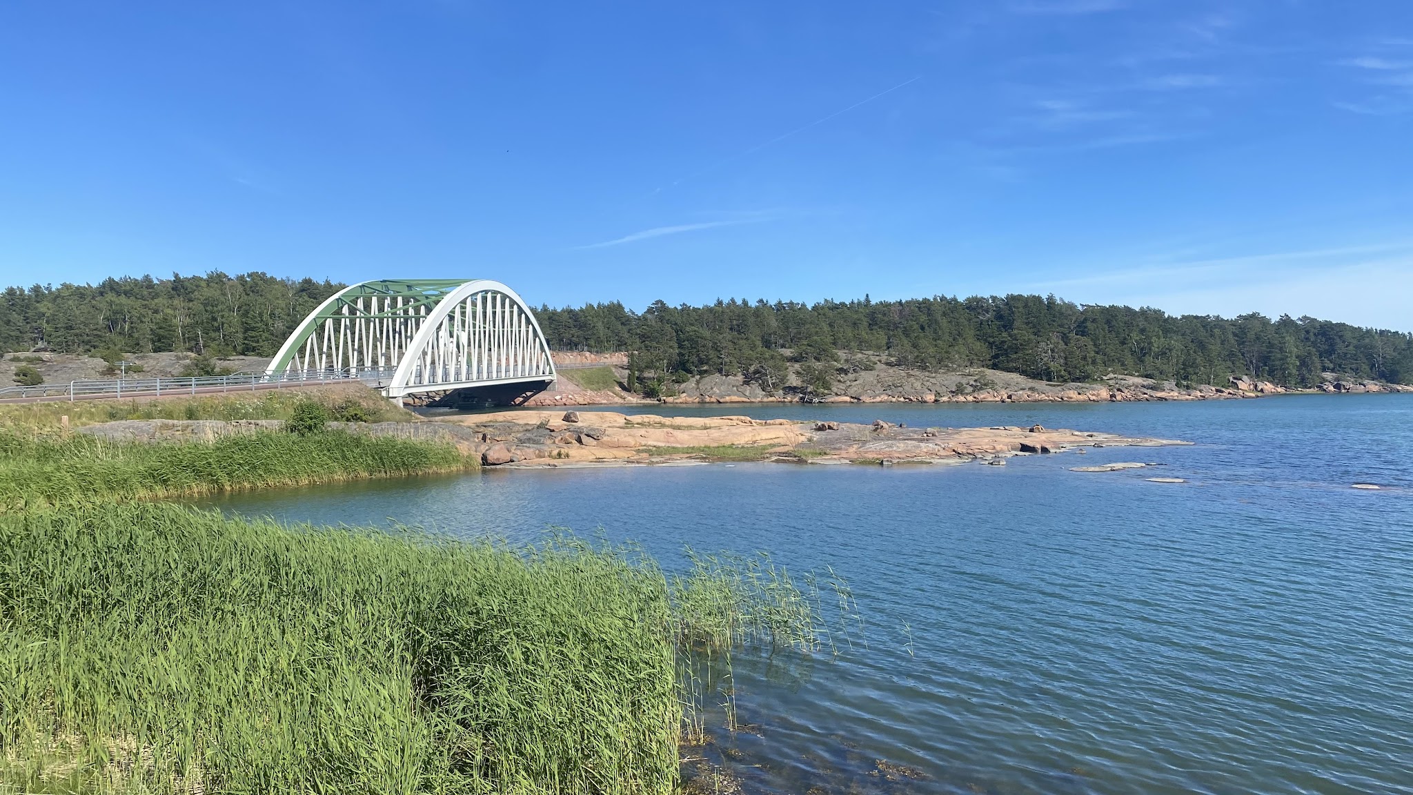 Bogenbrücke über blaues Küstenwasser, umgeben von Felsen, Schilf im Vordergrund und Nadelwald unter klarem Himmel