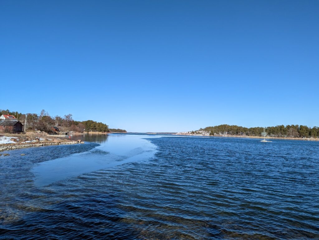 Weitläufige Bucht unter klarem Himmel; tiefblaues, teils vereistes Wasser, Ufer mit Häusern und Kiefern.