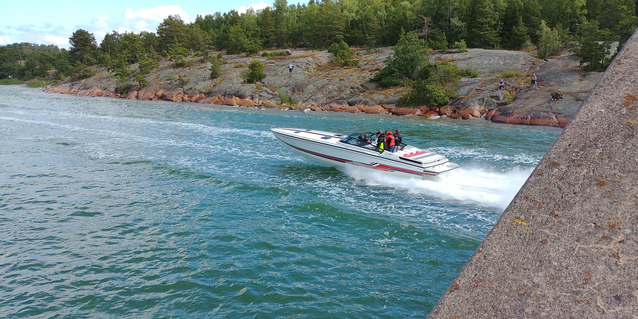 Schnellboot zieht weiße Gischt über türkisblaues Küstenwasser; felsige, bewaldete Uferkulisse im Hintergrund.