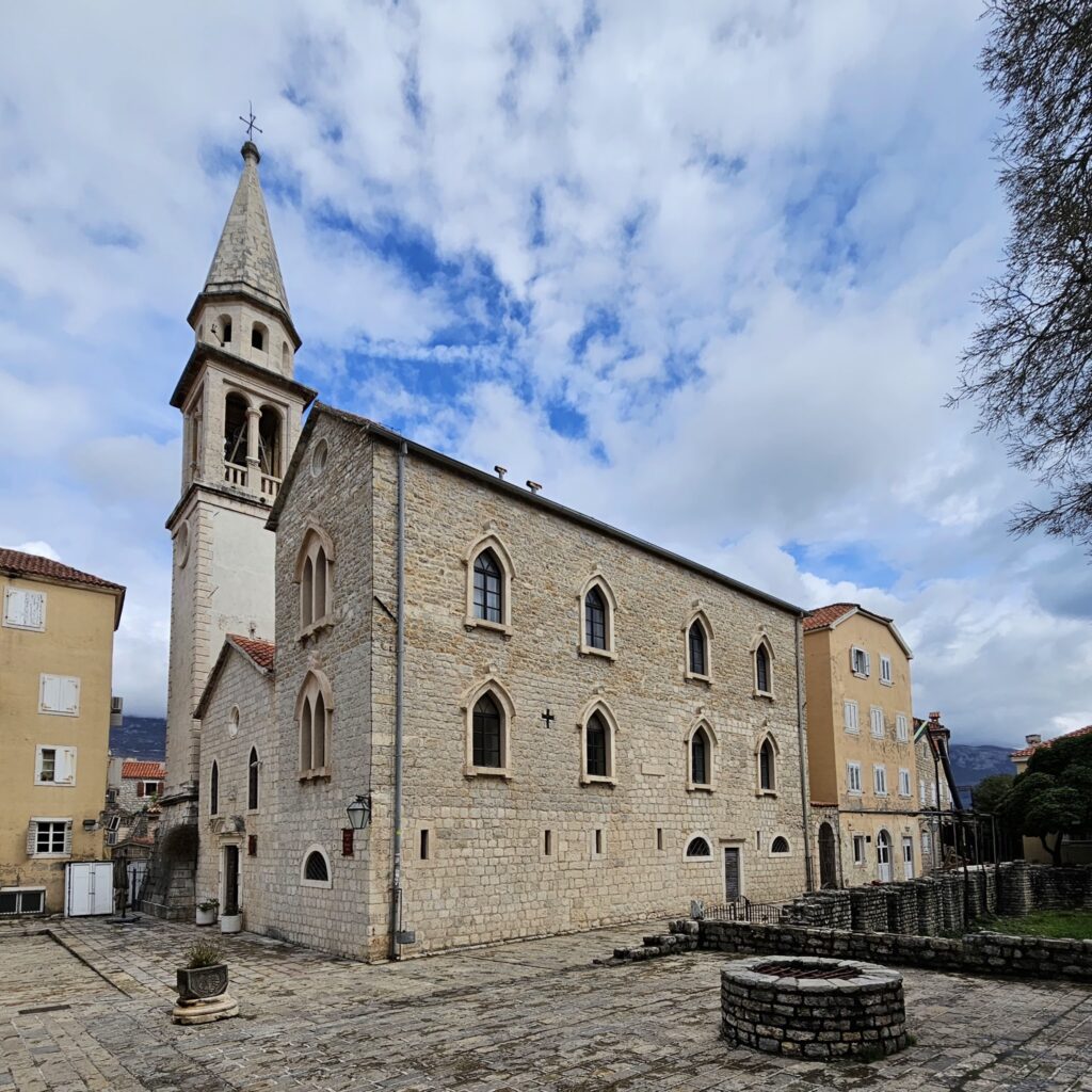 Romanische Steinkirche mit spitzem Glockenturm an gepflastertem Platz unter teils blauem, wolkigem Himmel