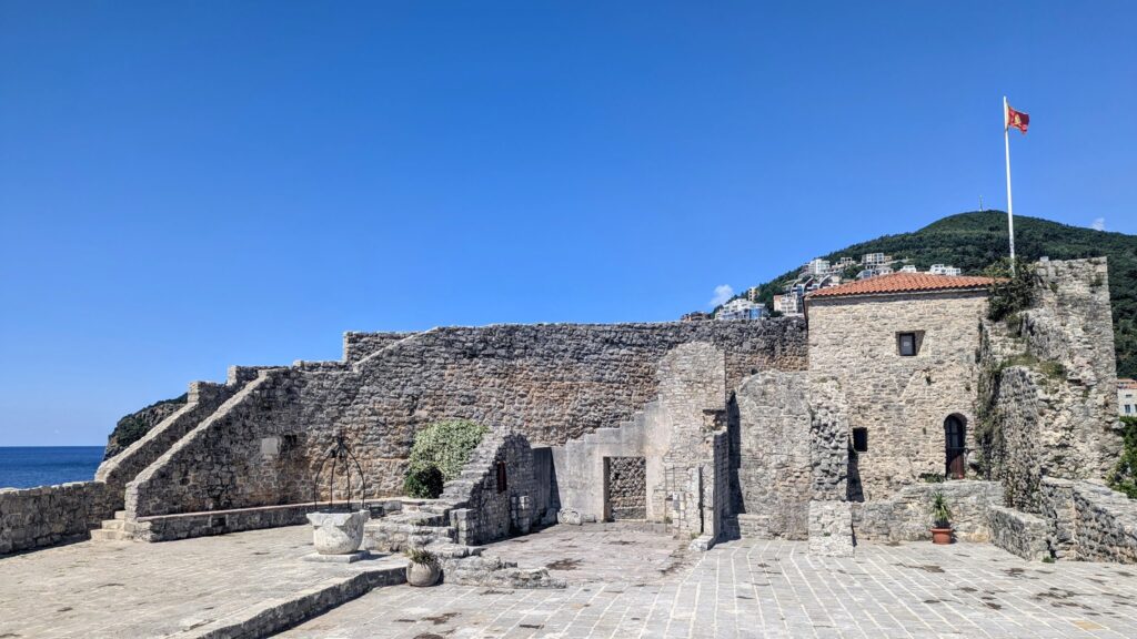 Steinerne Festungsmauern am Meer, Innenhof mit Brunnen; rote Flagge weht vor grünem Hügel unter klarem Himmel.