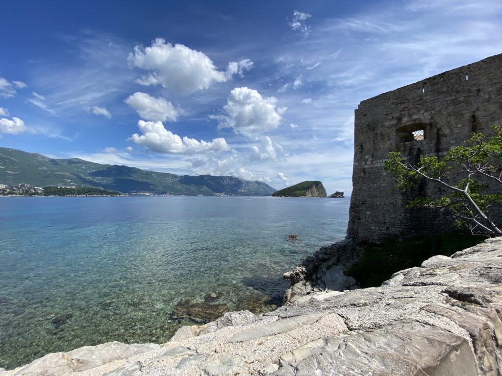 Klares türkisfarbenes Meer vor fernen Bergen und kleiner Insel; rechts steht eine alte Steinmauer einer Festung.