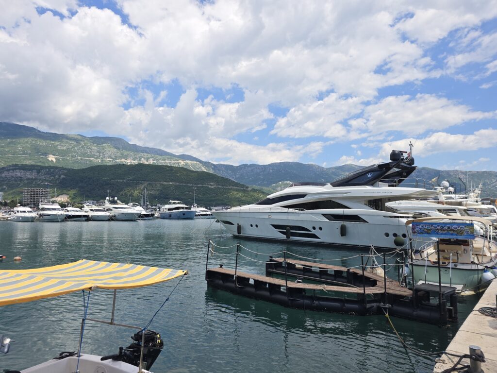 Luxusyacht am Steg, weitere Boote im klaren Hafen vor grünen Bergen und teils bewölktem Himmel.