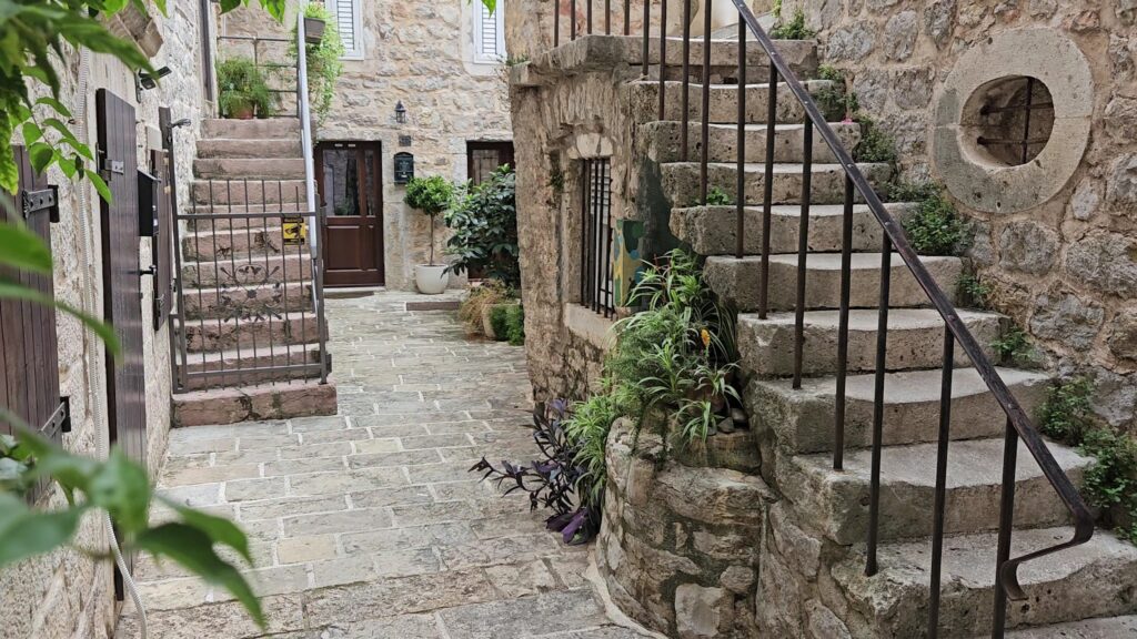 Schmale steingepflasterte Gasse mit zwei Außentreppen, Holztüren und vielen Topfpflanzen