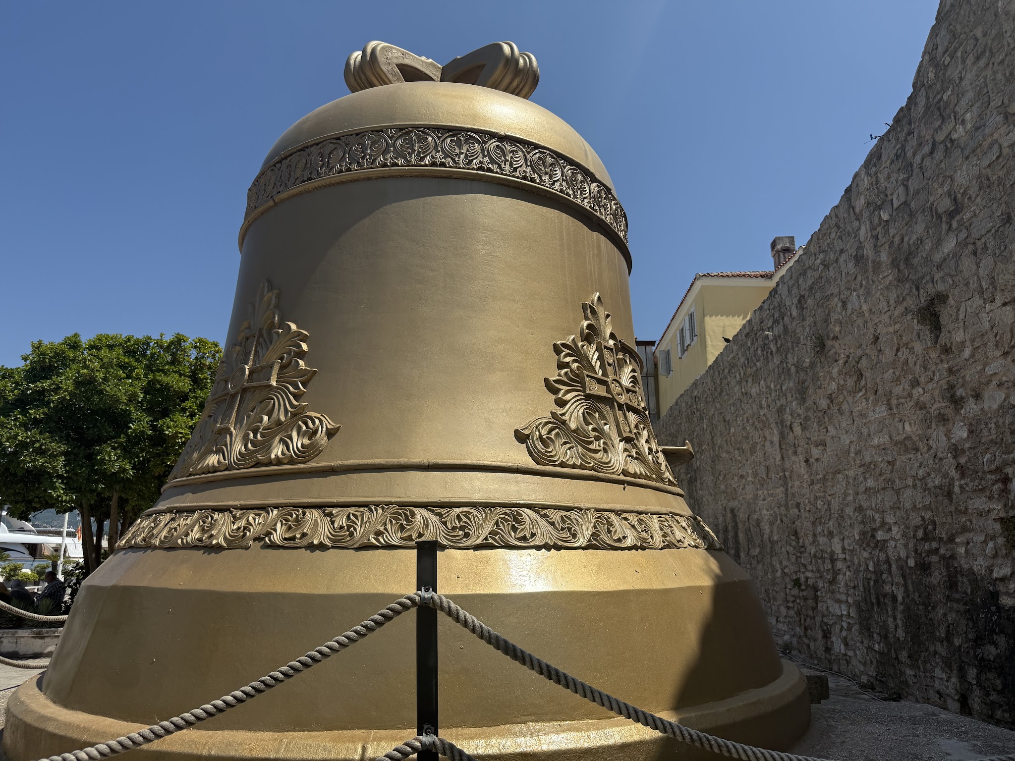 Überdimensionale goldene Glocke mit ornamentalen Reliefs, von Seilen abgesperrt, vor Steinmauer unter blauem Himmel.