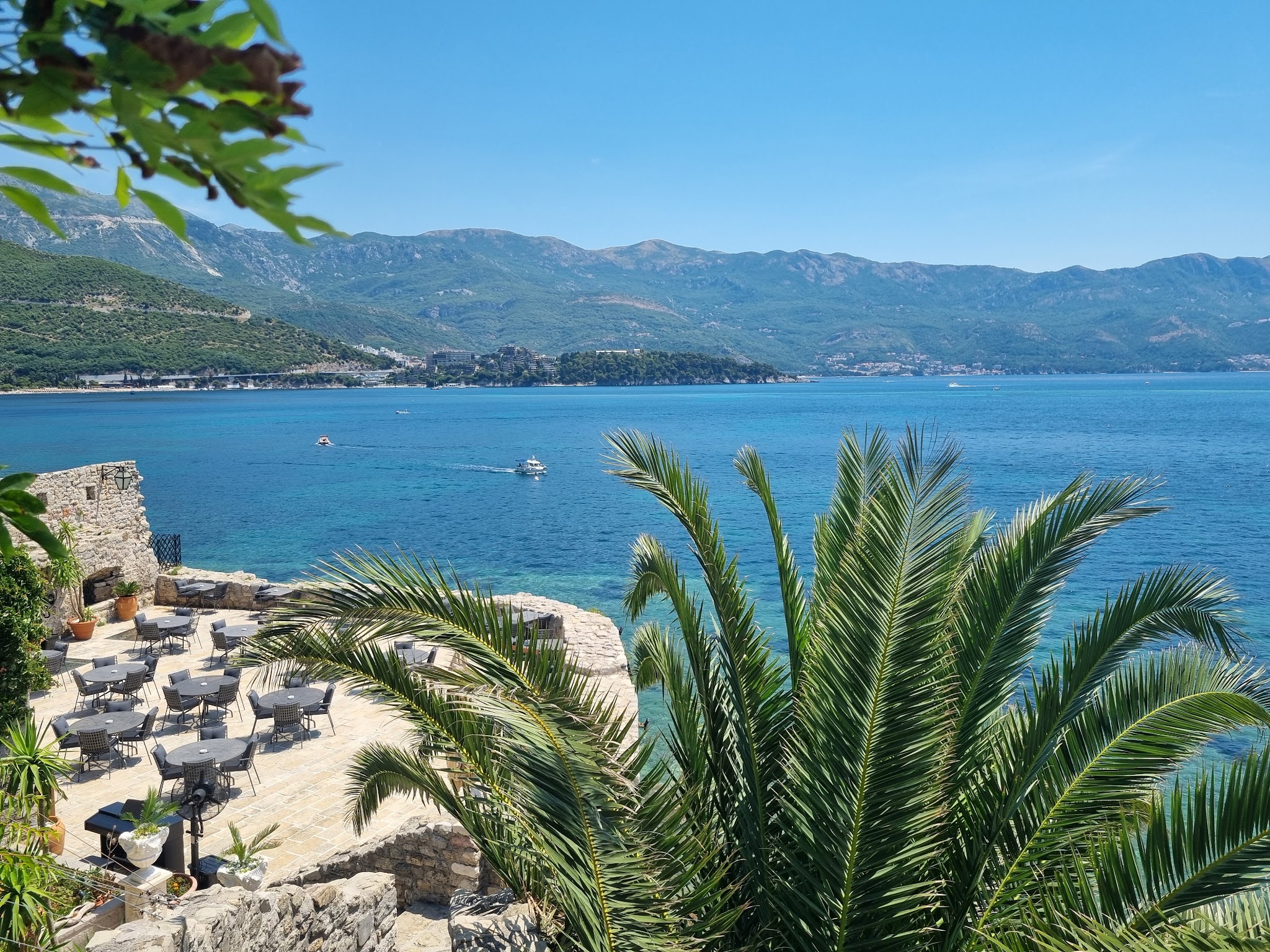 Terrasse mit leeren Café-Tischen unter Palmen, Blick auf türkisblaues Meer und grüne Küstenberge.