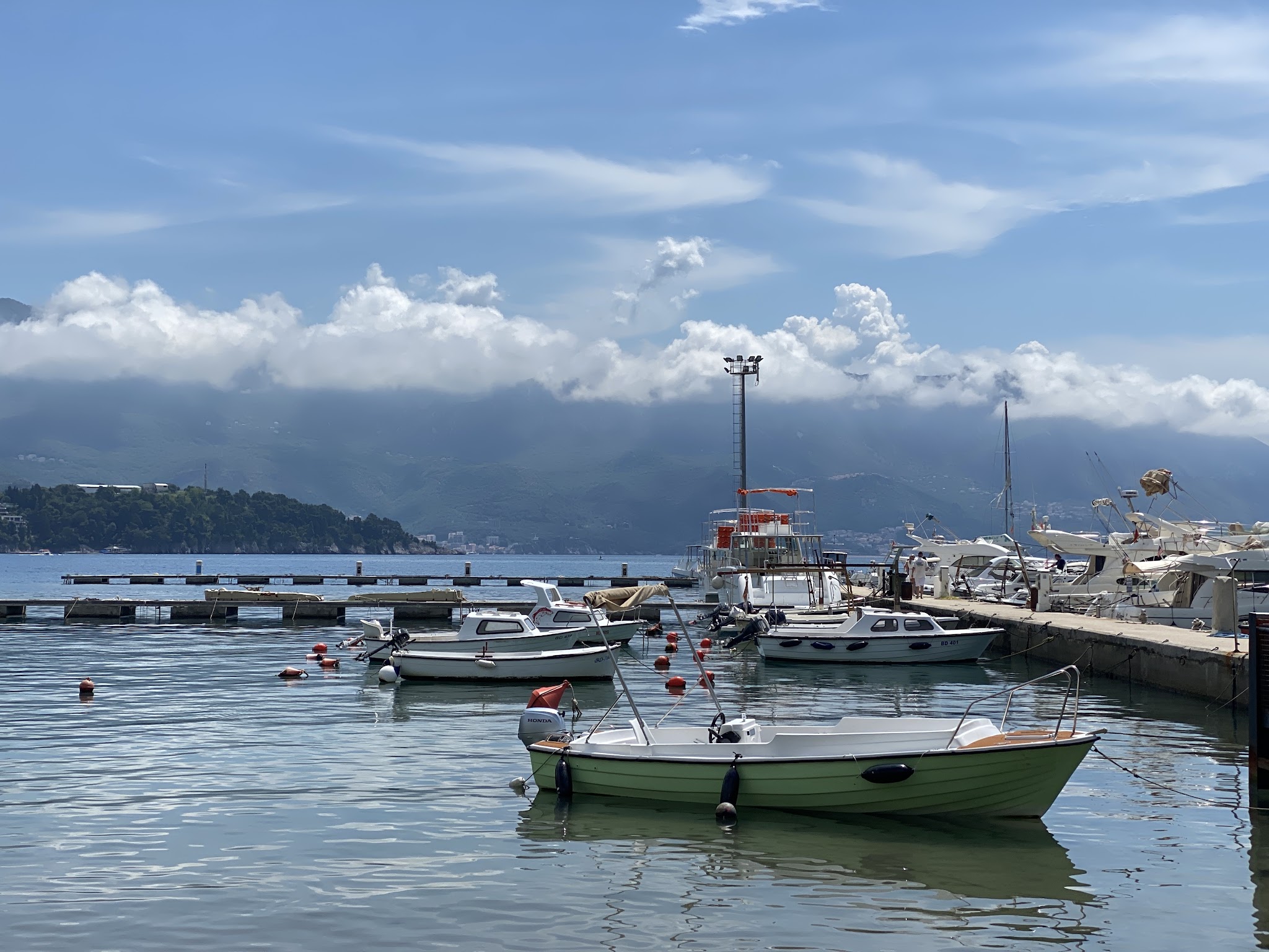 Ruhiger Yachthafen; kleine Boote im Wasser, dahinter wolkenverhangene Berge unter blauem Himmel.